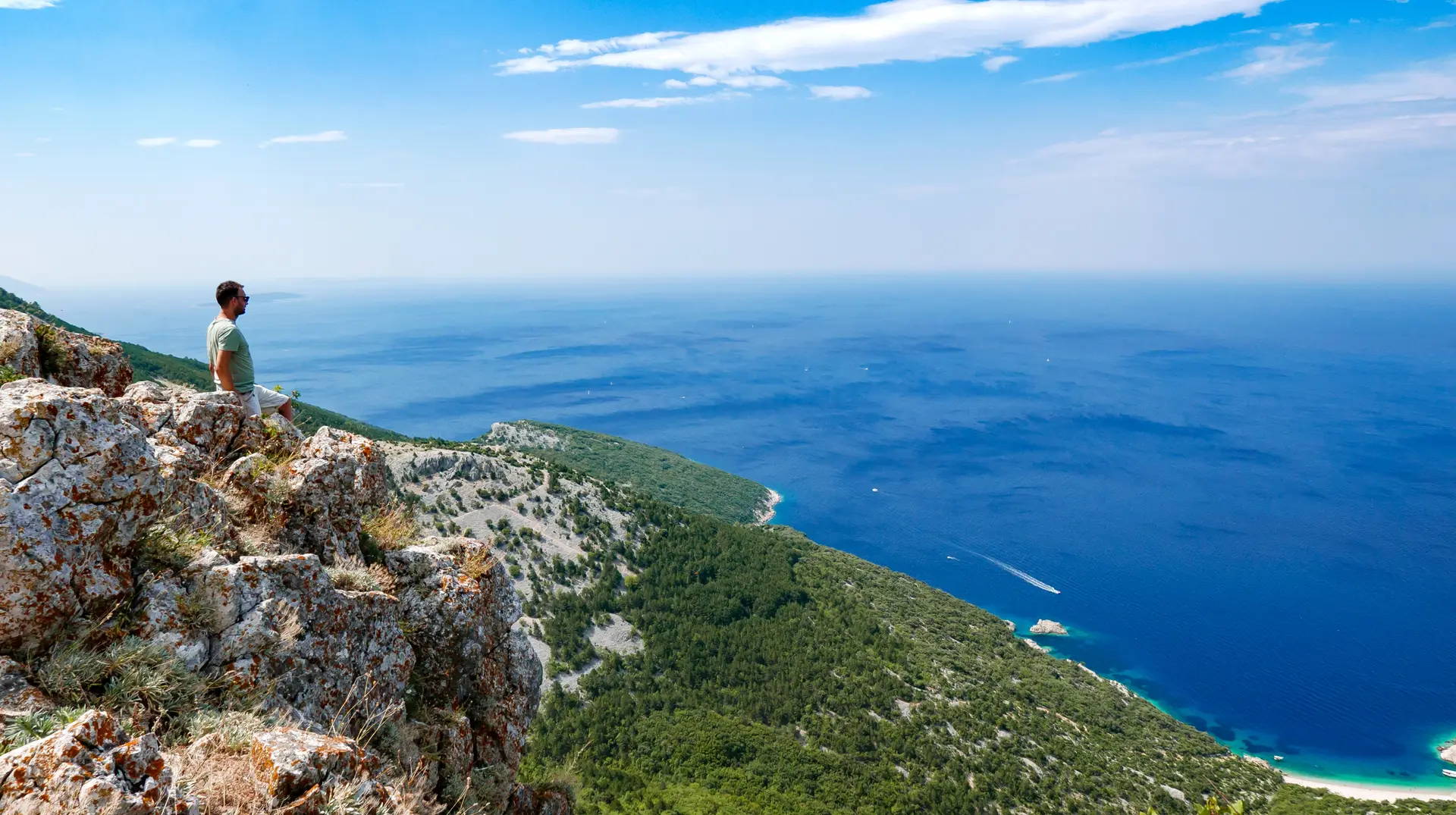 Young man standing on the edge of a cliff high above the sea looking at amazing adriatic coastline.